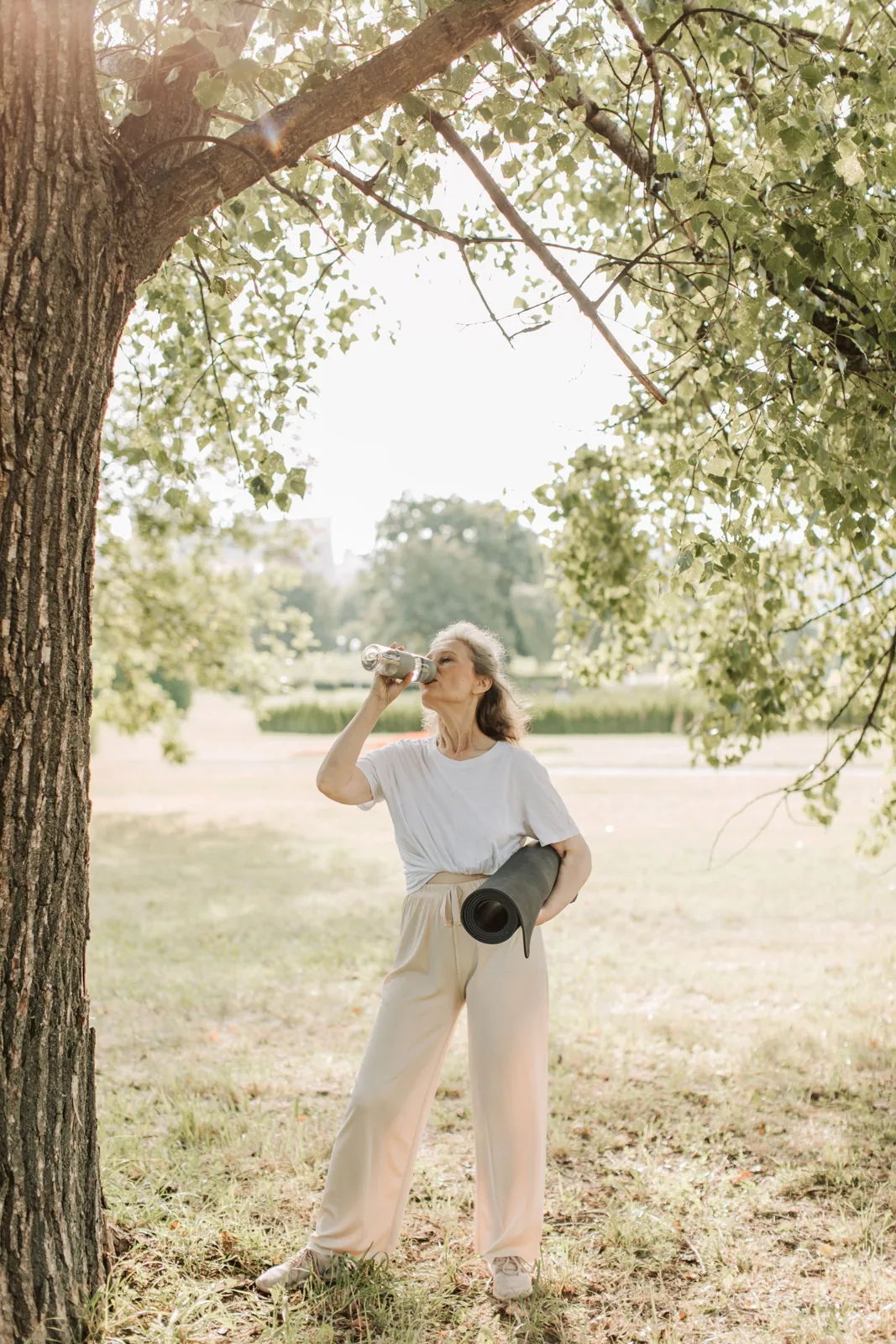 An older woman carrying a yoga mat drinks water beneath a sunlit tree in a park