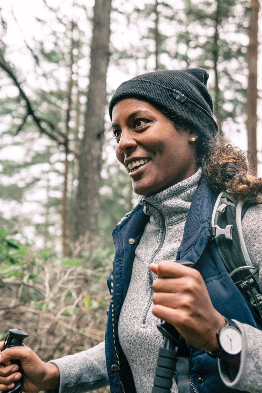 A smiling woman wearing a knit hat and backpack holds trekking poles on a forest walk