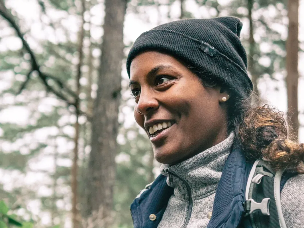 A smiling woman wearing a knit hat and backpack holds trekking poles on a forest walk