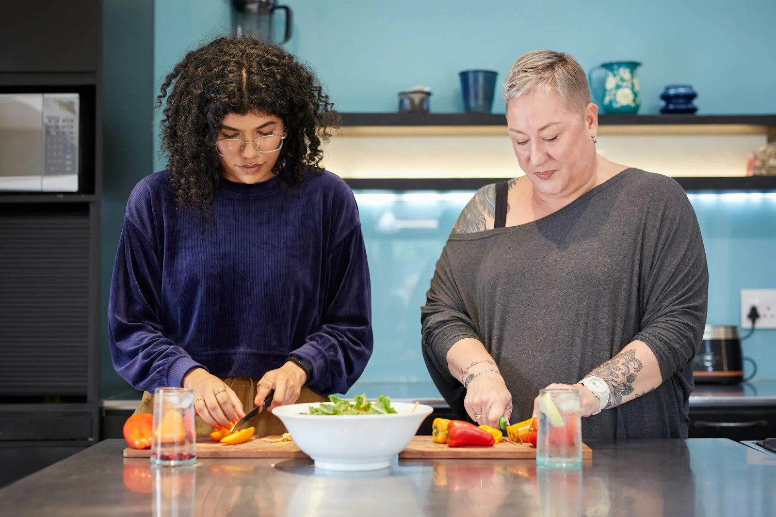 Two women prepare peppers and leafy vegetables together at a kitchen counter